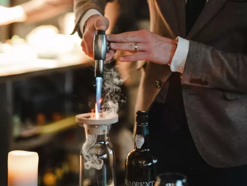 A person in a suit uses a torch to create a smoky effect in a glass decanter, with a bottle of liquor nearby.