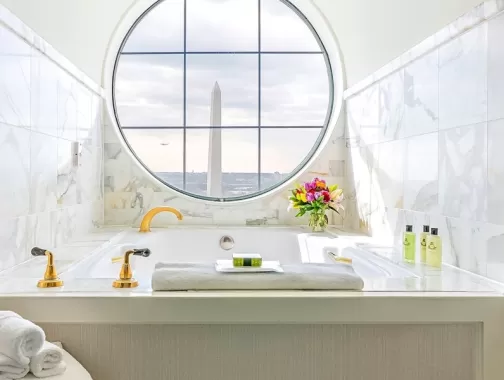 A luxurious bathroom with a marble bathtub and golden fixtures, featuring a round window with a view of the Washington Monument. The bathtub has towels, flowers, and toiletries arranged neatly.