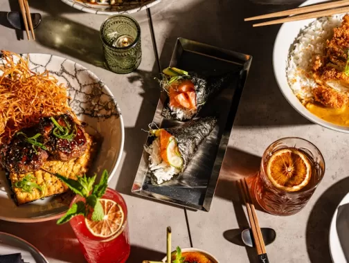 An overhead view of a diverse spread of Japanese dishes, featuring sushi rolls, rice bowls, and various appetizers, along with vibrant cocktails and meticulously plated dishes on a modern table setting.