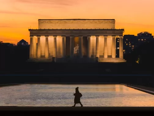 A silhouette of a person walking along the reflecting pool is set against the illuminated Lincoln Memorial at sunset, with a warm orange sky and city buildings in the background.