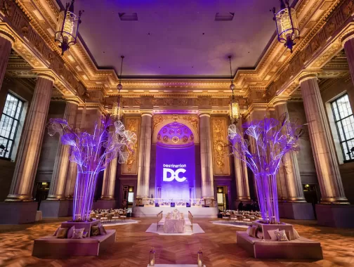 A grand event space with tall columns and ornate architecture, illuminated by purple lighting. The backdrop features a prominent "Destination DC" logo. Stylish seating areas with plush cushions are arranged in the foreground, complemented by artistic, tree-like sculptures.