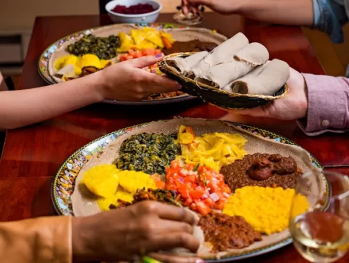 A vibrant Ethiopian meal served on large platters with colorful dishes, accompanied by injera bread, as diners share food and enjoy wine.