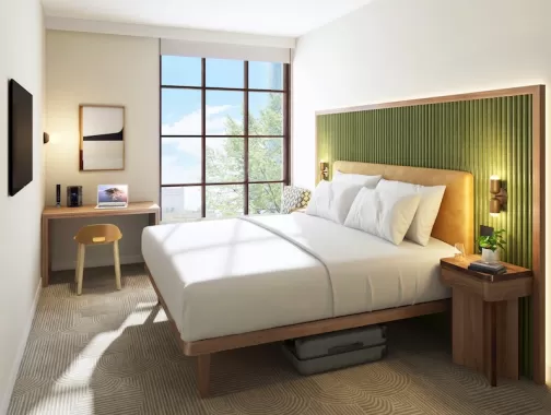 A modern hotel room with a green-accented headboard, large windows and a minimalist workspace.