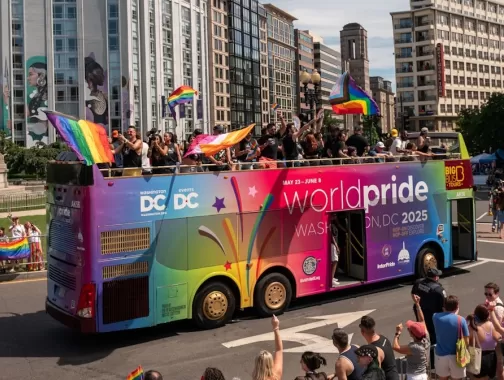 A colorful double-decker bus promoting WorldPride Washington, DC 2025 drives through a lively parade with waving rainbow flags and cheering crowds.