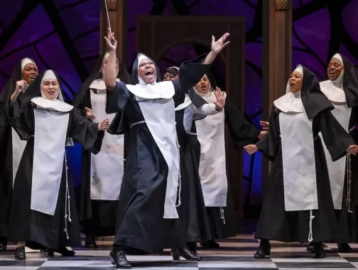 A nun holding a conductor ’ s wand dances animatedly with arms stretched up in the air as a large group of nuns dance and sing behind her.