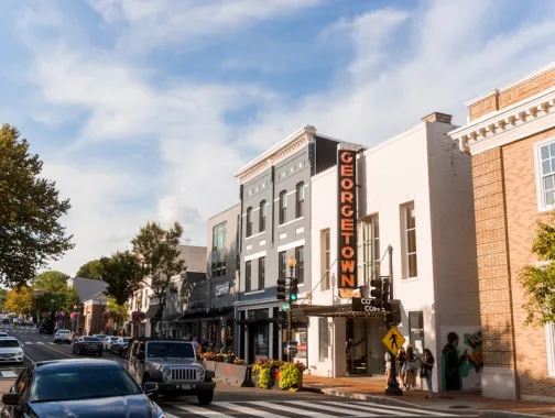 Pedestrians and cars pass by storefronts under a bright Georgetown sign on a bustling street in the historic Georgetown neighborhood of Washington, DC.