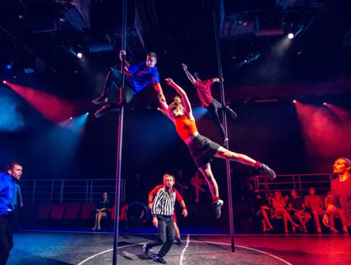 Acrobats perform daring moves on poles while a referee and audience watch during a high-energy scene from Duel Reality.