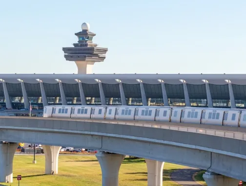 Silver Line Metro train traveling on elevated tracks with Washington Dulles International Airport terminal and air traffic control tower in the background.