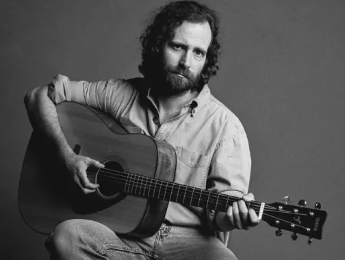 Comedian and musician Kyle Mooney poses with an acoustic guitar in a black and white promotional portrait.