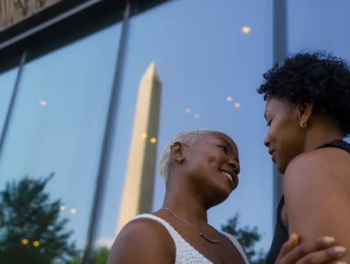 A couple shares an intimate moment in front of the Smithsonian National Museum of African American History and Culture, with the Washington Monument reflected in the glass.