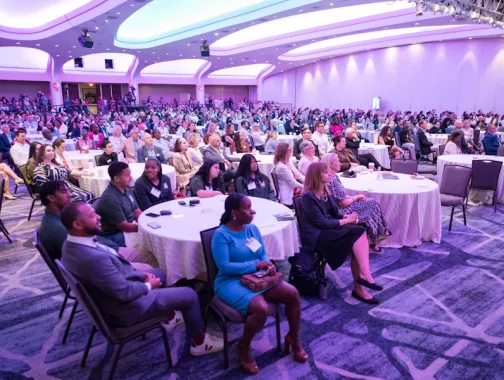 A large audience sits at round tables in a purple-lit conference hall, attentively listening to a presentation.