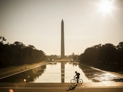 A cyclist rides past the Reflecting Pool with the Washington Monument rising in the background at sunrise.
