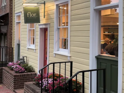The exterior of Flor Coffee + Books, a quaint home painted yellow with inviting flowers and a red door. 