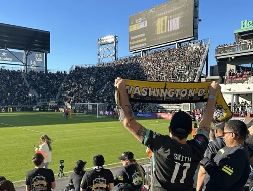 A Washington Spirit fan holds up a yellow scarf reading “Washington, DC” in a packed Audi Field stadium during a soccer match.
