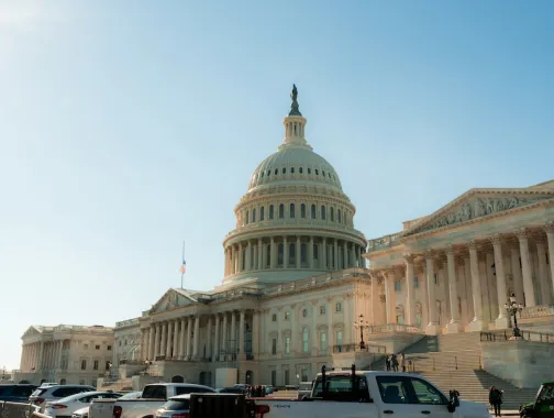 Cars park along the street in front of the U.S. Capitol as warm late-afternoon sunlight illuminates the dome and columns.