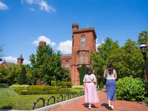 Two women walk along a garden path toward the Smithsonian Castle surrounded by lush greenery.
