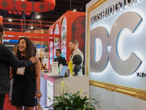 Attendees gather around a Washington, DC destination booth featuring illuminated signage and large digital displays at a travel trade show.