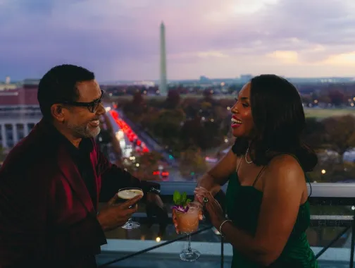 A couple enjoys cocktails on a rooftop terrace with the Washington Monument visible in the background at dusk.