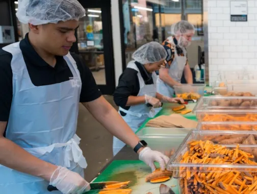 Three people wearing hairnets, gloves, and aprons slice sweet potatoes at a food preparation station with trays of cut vegetables.