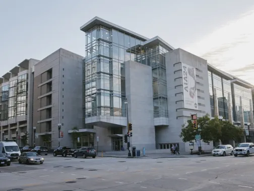 Un moderno edificio museale in vetro e cemento, situato all'angolo di una strada cittadina, con auto e pedoni sotto un cielo parzialmente nuvoloso.