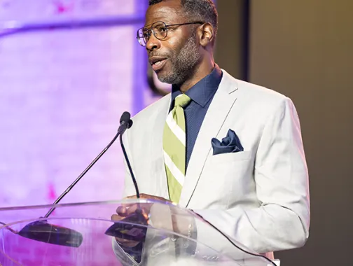 Elliott L. Ferguson, II in light suit speaking at podium with microphone under purple lighting.