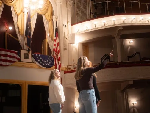 Two performers on stage beneath ornate theater balconies and chandelier.