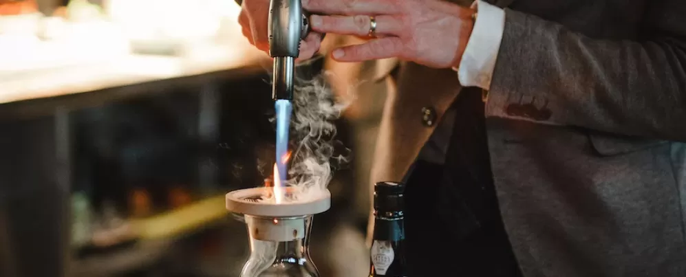 A person in a suit uses a torch to create a smoky effect in a glass decanter, with a bottle of liquor nearby.