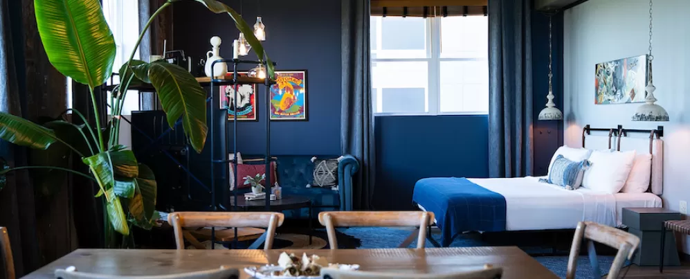 Cozy hotel suite with a dining table, large potted plant, blue sofa, and bed with blue bedding. Dark blue walls and framed artwork add color.