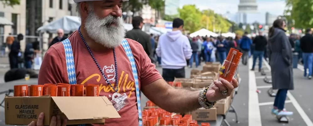 Um homem com barba branca, chapéu xadrez azul e suspensórios, distribuindo latas de cerveja laranja em um evento da Oktoberfest em Washington DC, com o edifício do Capitólio dos EUA visível ao fundo.