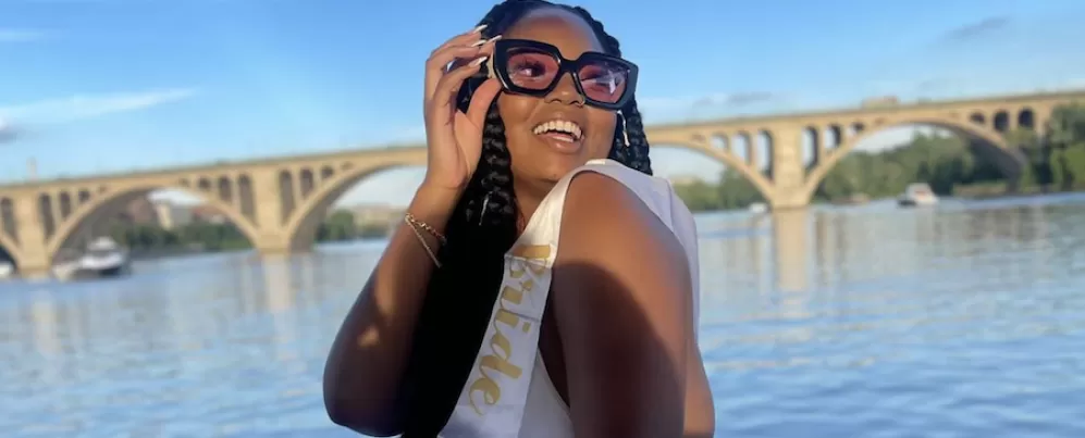 Eine lächelnde Frau mit einer großen Sonnenbrille und einer „Bride to Be“-Schärpe posiert neben einem Fluss mit einer Bogenbrücke im Hintergrund.