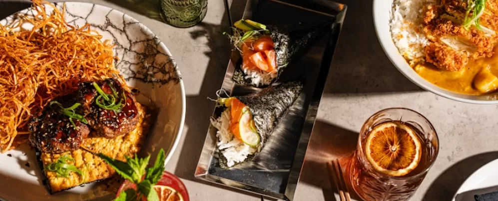 Uma visão aérea de uma variedade diversificada de pratos japoneses, com rolos de sushi, tigelas de arroz e vários aperitivos, além de coquetéis vibrantes e pratos meticulosamente preparados em uma mesa moderna.