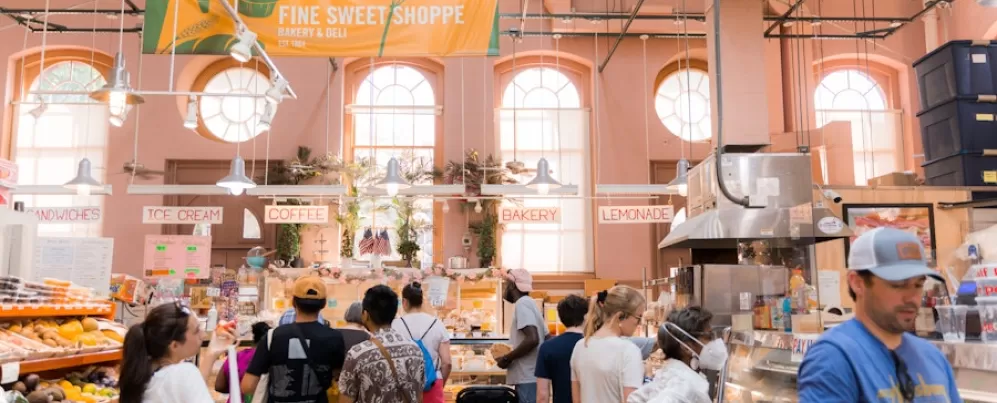 Het winkelend publiek staat in de rij bij een bakkerijkraam op de Eastern Market. De lichte en luchtige ruimte is gevuld met natuurlijk licht, met borden voor koffie, ijs en bakkerijproducten erboven.