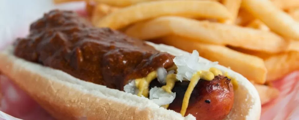 Esta imagem mostra um chili meio defumado do Ben's Chili Bowl, um clássico de DC que traz uma salsicha grelhada coberta com chili, mostarda e cebola, servida com batatas fritas.