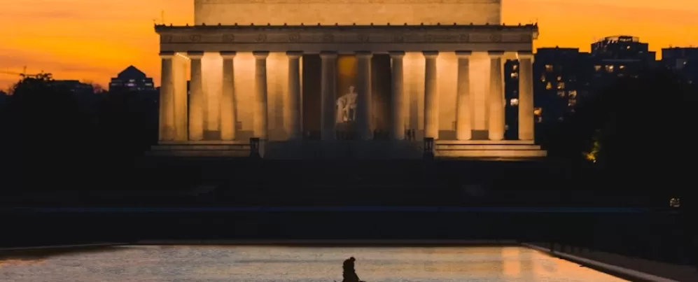 A silhueta de uma pessoa caminhando ao longo do espelho d'água contrasta com o iluminado Lincoln Memorial ao pôr do sol, com um céu laranja quente e prédios da cidade ao fundo.
