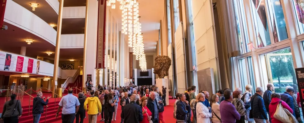 Un vestíbulo abarrotado dentro del Kennedy Center, con gente caminando junto a una gran escultura y candelabros bajo un techo alto.