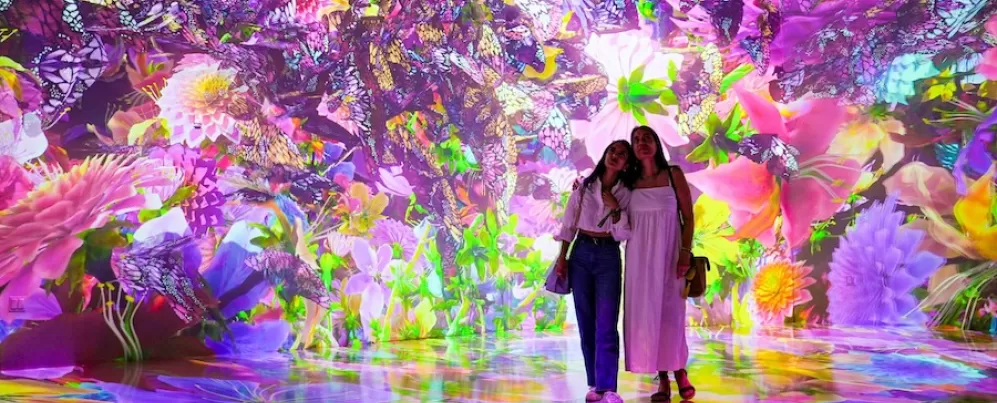 Dois visitantes estão em uma instalação de arte digital vibrante e imersiva na ARTECHOUSE, cercados por flores e borboletas coloridas e brilhantes.
