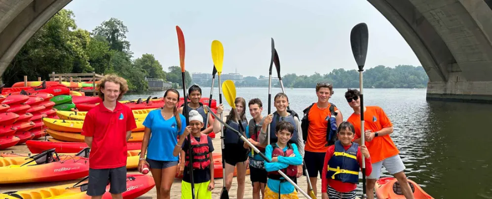 Group of kids and instructors with paddles posing on a dock surrounded by colorful kayaks under a bridge.