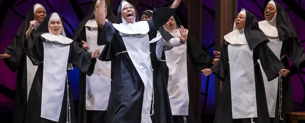 Une religieuse tenant la baguette d'un chef d'orchestre danse avec animation, les bras tendus en l'air, tandis qu'un grand groupe de religieuses dansent et chantent derrière elle.