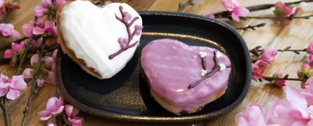 Dois donuts em formato de coração decorados com ramos de flores de cerejeira estão sobre uma mesa enfeitada com flores de cerejeira.
