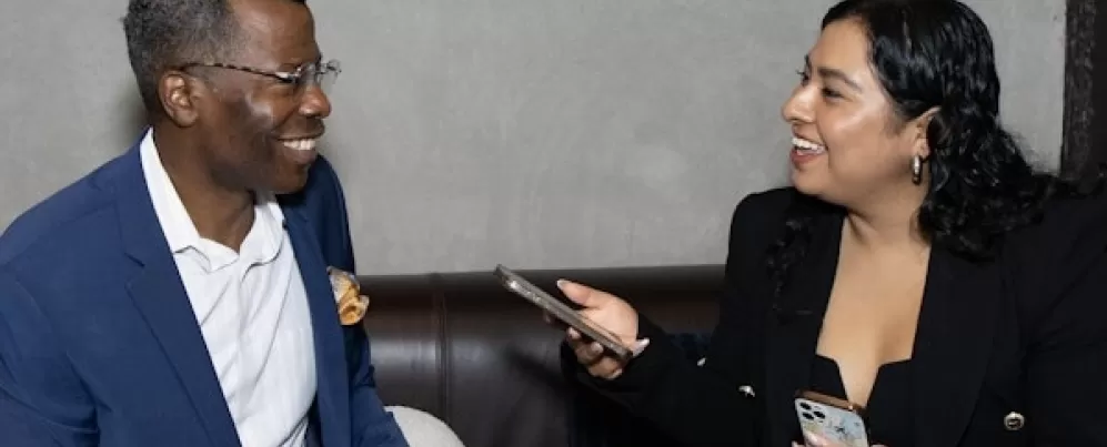 Cette image montre deux personnes assises sur un canapé, engagées dans une conversation amicale et professionnelle. La femme de droite tient un téléphone, réalisant peut-être une interview. Toutes deux sourient, suggérant une discussion positive et engageante. Le cadre paraît décontracté mais professionnel, peut-être un événement médiatique ou de réseautage.