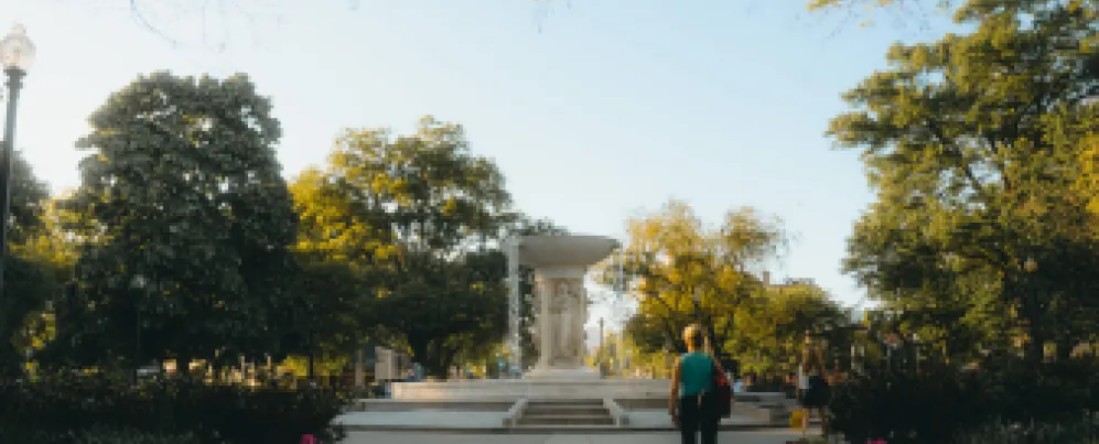 A scenic view of Dupont Circle featuring the central fountain, blooming flowers, and people strolling along the sunlit pathways.