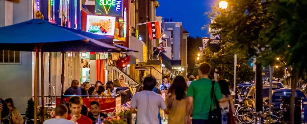 People walk down a street lit up with neon signs and busy restaurants and bars. 