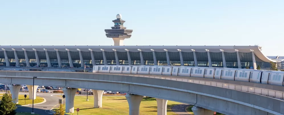 Der Metrozug der Silver Line fährt auf Hochgleisen mit dem Terminal des Washington Dulles International Airport und dem Flugsicherungsturm im Hintergrund.