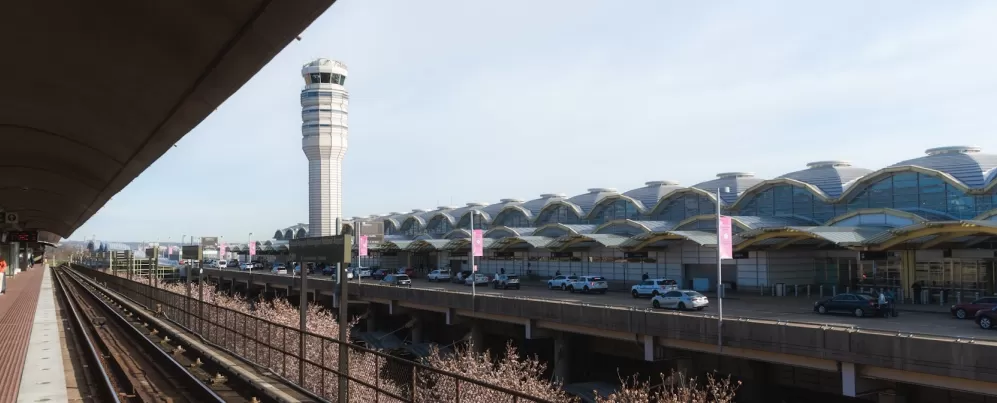 Blick auf die Außenseite des Ronald Reagan Washington National Airport mit dem Flugsicherungsturm und den U-Bahn-Gleisen im Vordergrund an einem klaren Tag.