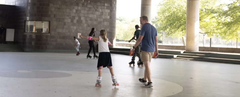 A father walks beside his daughter while she rollerblades.