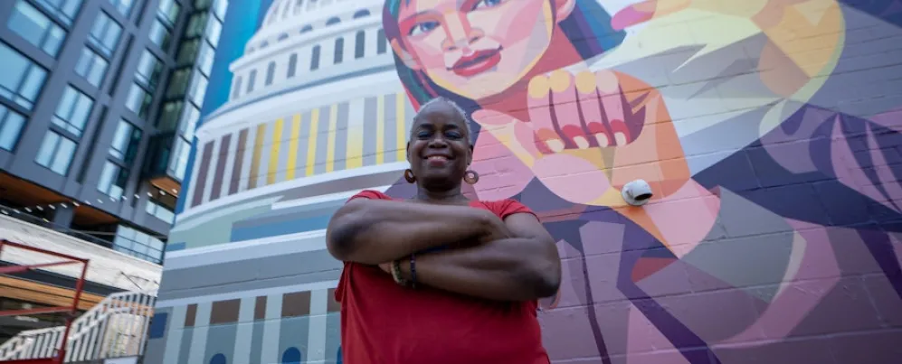 A person crosses their arms and smiles for a portrait in front of a colorful mural that depicts someone signing with the U.S. Capitol in the background. 
