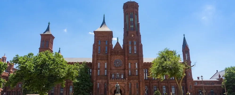 Visitors walk across the National Mall toward the red sandstone Smithsonian Castle on a sunny day.