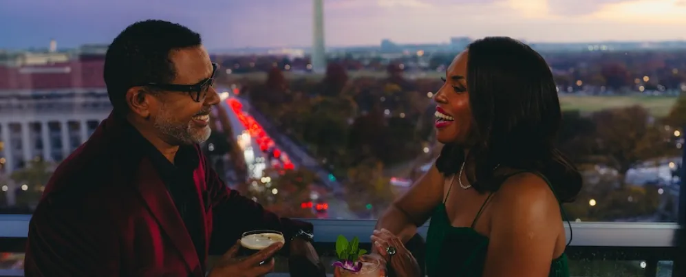 Um casal desfruta de coquetéis em um terraço na cobertura, com o Monumento a Washington visível ao fundo ao entardecer.