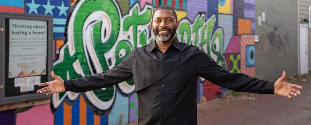 A smiling man stands with his arms open in front of a colorful street mural in Washington, DC.