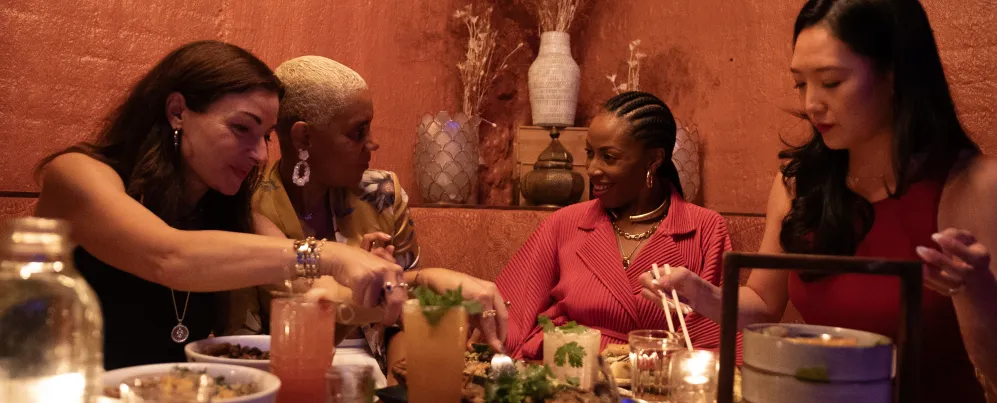 A group of friends share small plates and colorful cocktails at Doi Moi in Washington, DC, seated around a candlelit table with warm, textured walls in the background.